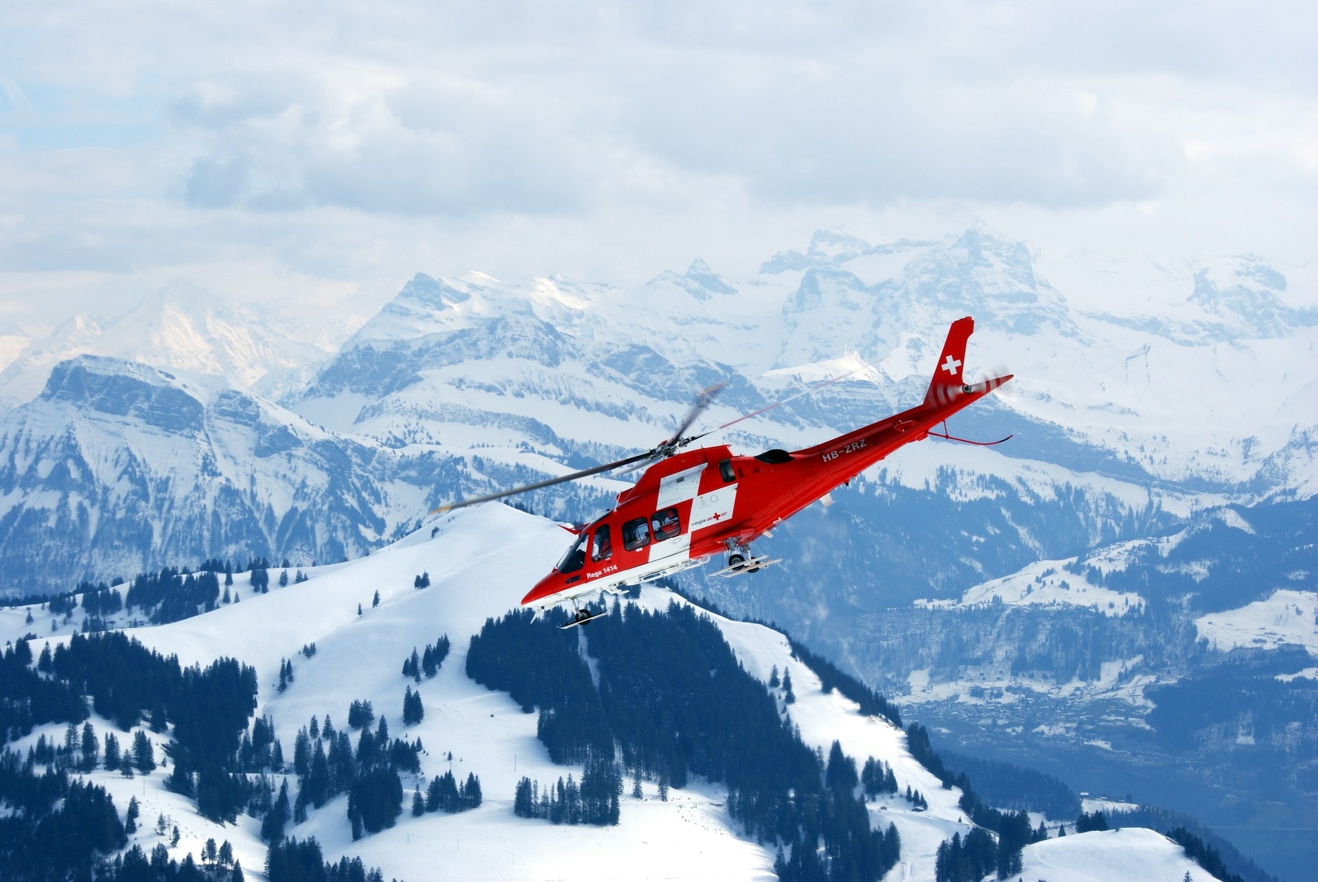 Red helicopter flying over snow-covered alpine mountains and forests